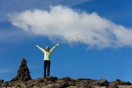 一位美丽的年轻女子站在地平线上,高举双臂庆祝登上山顶,她旁边是一块