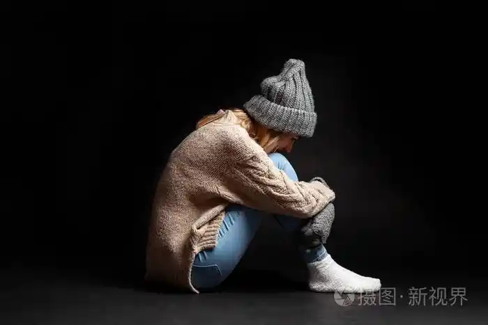 女孩独自坐在地板上,黑色的空虚背景,用手拥抱她的腿,把她的脸垂在